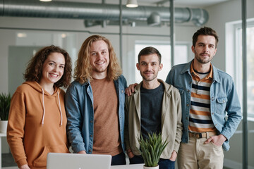 Group of young adult Caucasian men and woman standing together smiling in modern office environment, looking at camera, showing teamwork and collaboration, indoor setting