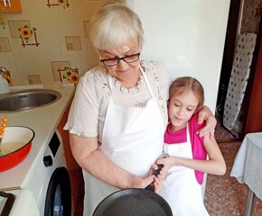 A grandmother and her granddaughter in white aprons are making pancakes in the kitchen.