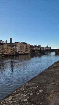 Vertical panning shot along the Arno River in Florence, Italy, starting at Ponte Santa Trinita and ending at the iconic Ponte Vecchio on a clear day.