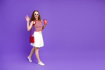 Young woman with red hair wearing a striped top and white skirt waves and smiles while holding a coffee cup against a purple background