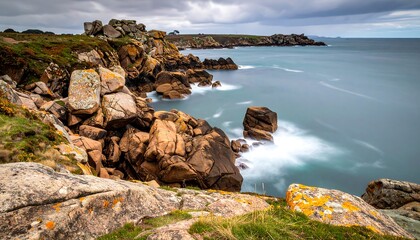 Rugged coastal scene with rocky cliffs meeting blurred ocean under a cloudy sky, taken from a grassy foreground