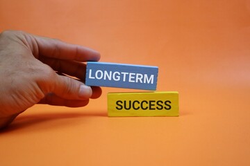A hand places a blue block labeled LONG TERM over a yellow block labeled SUCCESS, illustrating long-term success, strategic growth, and goal achievement in a orange background.
