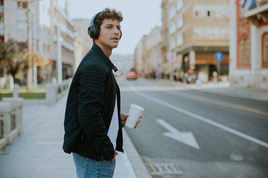 Man wearing headphones holding a coffee cup stands on a city sidewalk