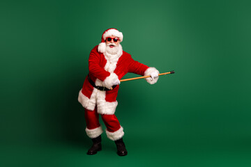 Santa in red suit with white fur hat holding a baton smiles in a green studio backdrop