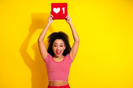 Cheerful young woman holding a red square sign with a white heart icon and number one against a yellow background