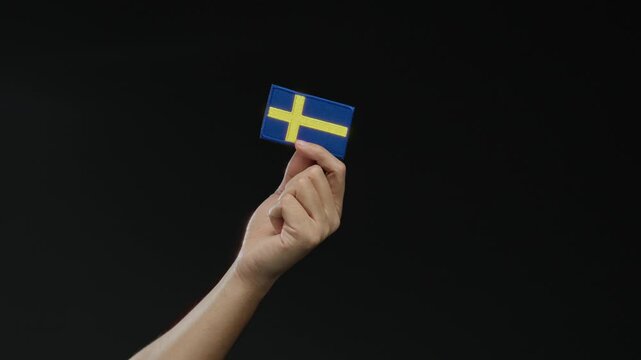Hand holding a swedish flag patch against a black background, symbolizing swedish nationality and pride, isolated and prominent in the scene, capturing cultural essence and identity.