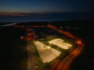 Aerial nighttime view of coastal skatepark bright concrete ramps and bowls glow amid dark surroundings, curved roads, tents, and distant waterline frame this illuminated recreational hub.