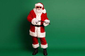 Santa in a bright red suit with fluffy white trim and belt poses with props in front of a solid green background for festive holiday promotion