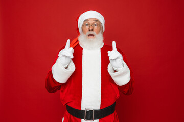 Funny happy excited old bearded Santa Claus face wearing costume looking at camera showing pointing fingers up advertising Christmas promotion, New Year xmas discount ad isolated on red background.