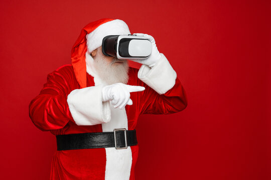 Santa Claus in black virtual reality glasses makes gestures with his hands. Surprise, emotion. New technology. Isolated on red background. Merry Christmas New Year concept.