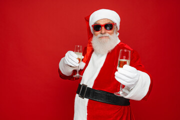 Photo of retired old man grey beard funky holding two full glasses golden wine having fun wear santa x-mas costume suspender sunglass cap isolated red color background. Merry Christmas New Year