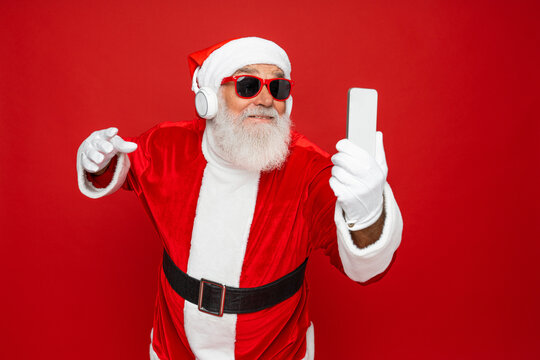 Crazy santa claus with grey beard wireless modern headset headphone holding smart phone looking at mobile screen listen music dance wear sunglass cap isolated bright shine red color background