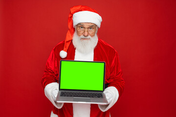 Smiling Santa Claus man in Christmas hat red coat holding using laptop pc computer showing demonstrate blank empty screen isolated on red background. Happy New Year celebration merry holiday concept