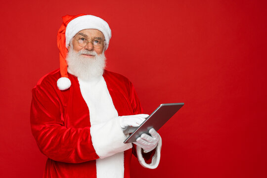 Elderly gray-haired Santa Claus man in Christmas hat red suit coat white gloves glasses working on tablet pc computer isolated on red background. Happy New Year celebration merry holiday concept