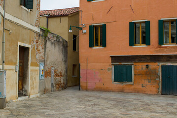 A quiet, stone-paved square sits nestled between aged buildings with distressed plaster in warm tones. Deep green shutters and doors provide striking contrast to the weathered walls. Venice, Italy.