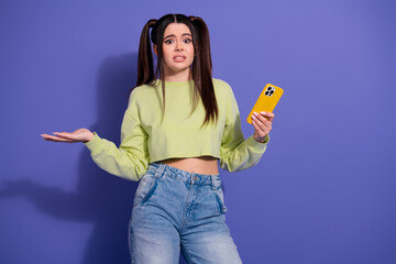 Young woman in casual attire holding a smartphone expressing confusion, standing against a purple...