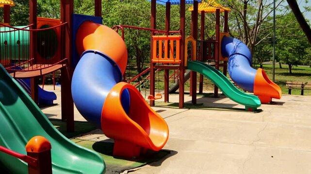 Colorful playground slides and tunnels under bright sunlight at a public park in Asuncion, Paraguay. Low angle panning shot showing vibrant structures and trees in the background.