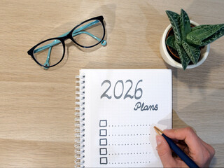 2026 Plans,  A spiral notebook on wooden desk with checklist for goals, planning for upcoming New Year., with eyeglasses, aloes and hand holding navy pen