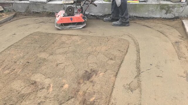 A worker operates a compactor to prepare a sand base for paver installation on a construction site. The focus is on achieving a smooth surface for laying bricks.