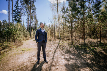 Obraz premium Man in formal suit stands confidently on a forest path, surrounded by tall trees and greenery, showcasing a blend of nature and elegance in a serene outdoor setting