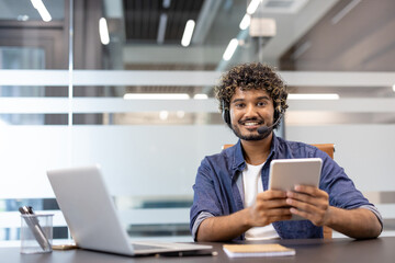 Portrait of a young Indian man in a headset sitting in an office at a table with a laptop, holding a tablet in his hand and looking at the camera with a smile