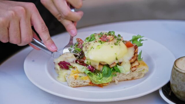 Hands cutting gourmet eggs benedict with fresh greens on a white plate in a cozy cafe setting.