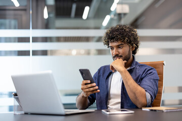 A young Muslim man sits in an office at a table with a laptop and looks thoughtfully at the screen of his mobile phone