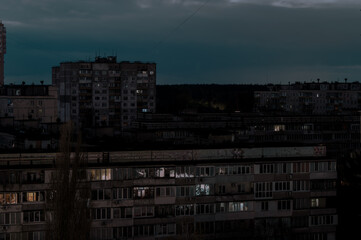 Fototapeta premium Distant view of a city during a dusk blackout with silhouetted buildings and faint lights visible in some windows under a dark, cloudy sky. 