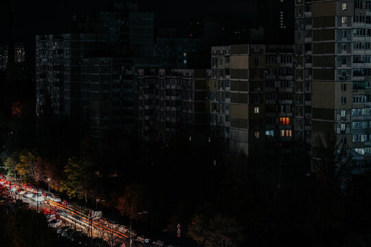 A darkened urban cityscape at night with minimal lights visible in apartment buildings and street traffic. The scene evokes a sense of blackout or power outage