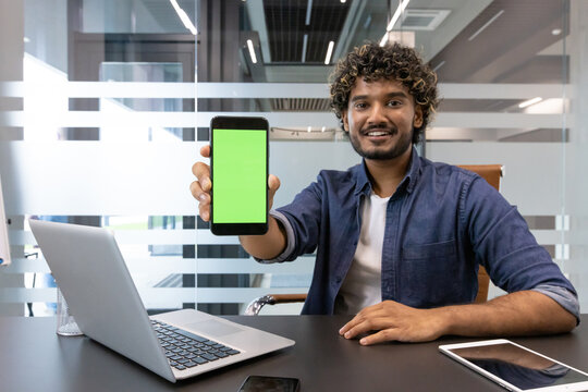 Portrait of a young Indian man sitting in an office at a desk and showing a phone with a green blank screen to the camera