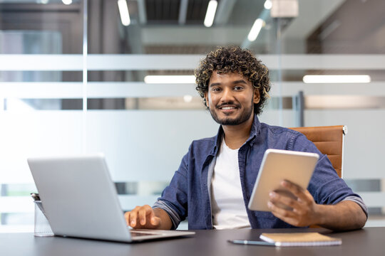 Portrait of a young Muslim man sitting at an office desk, holding a tablet in his hand and looking at the camera