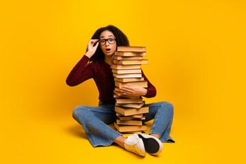 Young mixed race girl with glasses sits on a yellow background and stacks book while smiling...