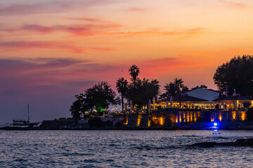 Sunset view of Side's historic peninsula with palm silhouettes against pink-orange sky, waterfront cafes illumination and tourist boat on evening sea. Side, Antalya, Turkey.

