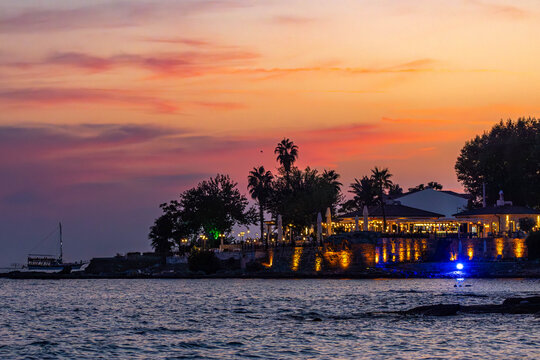 Sunset view of Side's historic peninsula with palm silhouettes against pink-orange sky, waterfront cafes illumination and tourist boat on evening sea. Side, Antalya, Turkey.
- Powered by Adobe
