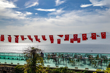 Cafe terrace with Turkish flag garlands and glass railings overlooking sparkling Mediterranean Sea, celebrating Republic Day with festive coastal atmosphere. Antalya, Turkey.

