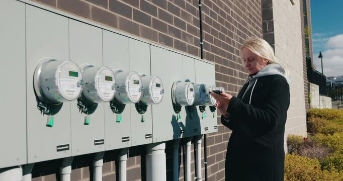 Woman checking outdoor electric meters and recording readings on her phone