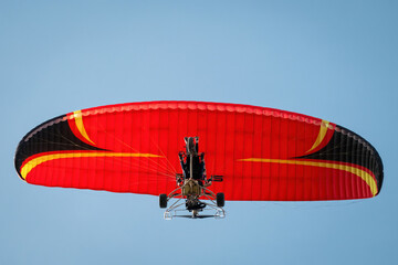 A powered paraglider with a bright red wing soars against a clear blue sky, representing freedom and adventure in flight.