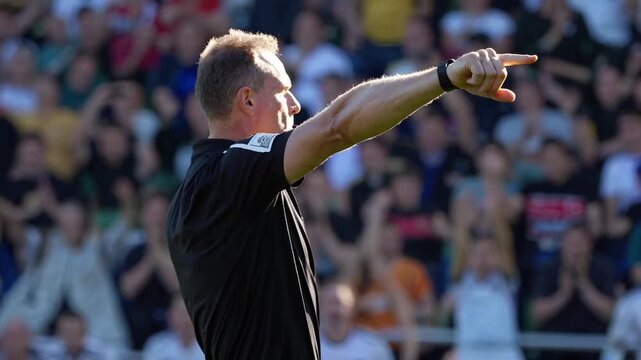 white male referee pointing during match, sunlit stadium, packed stands, extended arm signal, authoritative