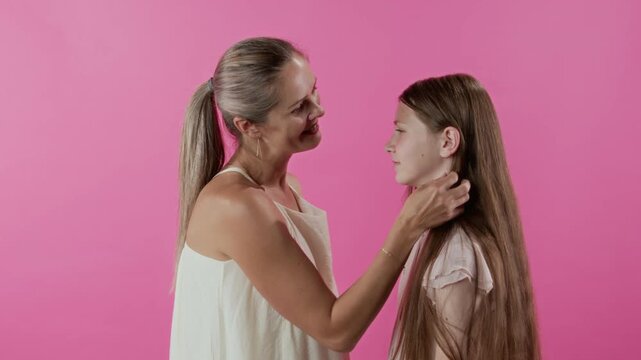 Portrait of adult woman gently touching hair of teenage daughter then two embracing expressing care and love in studio on pink backdrop, slow motion