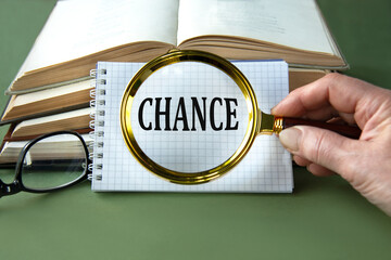 A woman's hand holds a magnifying glass over the word CHANCE on a green background with books