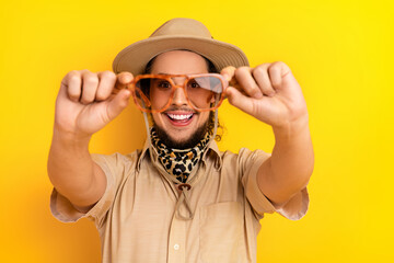 Young man in safari outfit holds orange sunglass toward camera smiling yellow background creating...