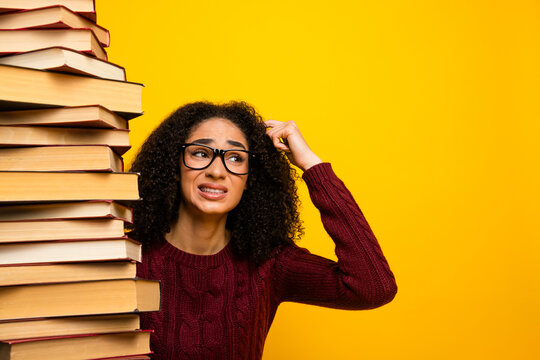 Young mixed race woman with glasses stands beside tall stack of book against yellow background showing stress while studying and learning