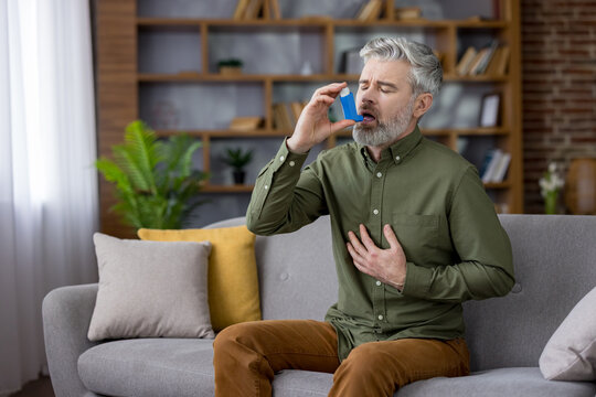 Mature man with white beard clutching his chest on a living room sofa during an asthma attack, using a blue inhaler for urgent breathing relief and medical treatment