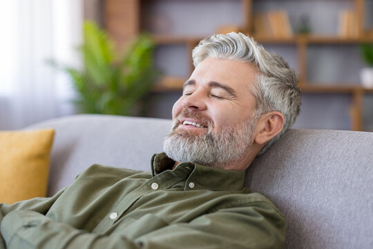 Mature man with grey hair and beard reclining on a cozy sofa at home, eyes closed and smiling, enjoying peaceful, stress-free leisure and calm wellbeing in the living room