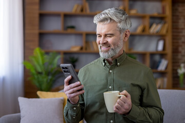 Mature bearded man smiling happily and holding a mobile phone while enjoying a hot drink on the sofa at home, connecting with friends and family online