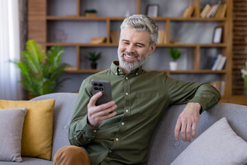 Grey-haired man relaxing on a comfortable sofa, smiling brightly and engaging with his smartphone, enjoying modern technology in a cozy home environment