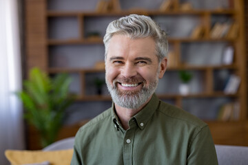 Smiling mature man with grey hair and beard, sitting at home in smart-casual shirt, confident and...