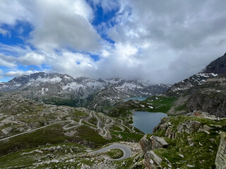 A dramatic wide-angle view captures the iconic Col del Nivolet Pass in the Italian Alps.The winding cycling road leads toward serene alpine lakes and a distant glacier, showcasing a majestic landscape