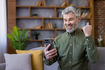 Mature man with a happy expression celebrating success and good news while looking at a smartphone, clenching his fist in an excited gesture, sitting on a sofa at home