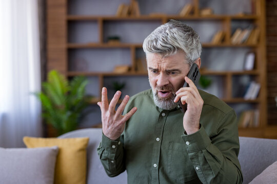 Senior man with gray hair talking on a mobile phone, experiencing stress and frustration during an emotional conversation or argument in his living room